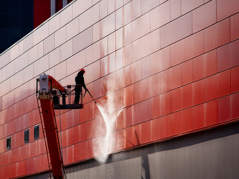 Worker wearing safety harness washes wall facade at height on modern building in a crane.