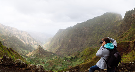 Fototapeta premium Traveler on the mountain edge making a photo of landscape. Deep clouds above green Xo-Xo Valley. Santo Antao Island, Cape Verde
