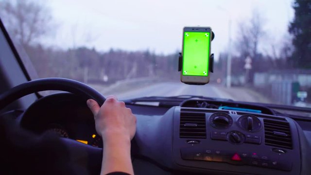 Someone driving a car at cloudy evening in country. Smartphone with a green screen in the holder on the windshield. Hand of a driver from time to time scrolling the smartphone.