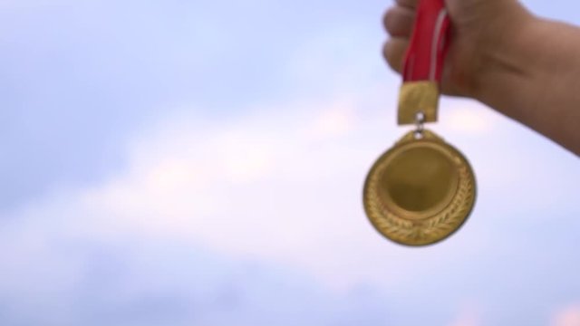 Winner hand raised and holding gold medals with ribbon against sunset light sky. Golden medal is awarded for highest achievement for sport or business. Success Awards concept, slow motion shot