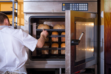 A male baker in white uniform and a beige apron bakes bread  from an industrial oven. Work at the...