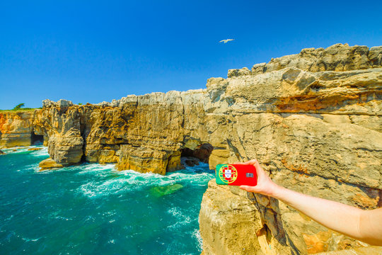 Hell's Mouth, Atlantic coast of Cascais, Portugal. Mobile phone with Portugal flag cover taking photos of Boca do Inferno a natural arch in rough cliff formation.Tourism and travel concept in Portugal