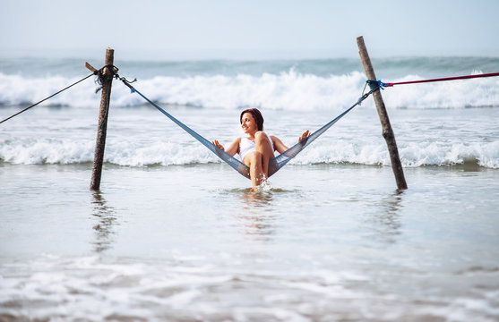 Woman in white swimsuit sits in hammock swing over the ocean waves