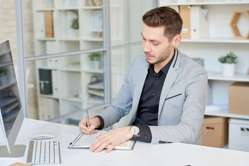 Portrait shot of confident young manager taking notes while brainstorming on start-up project, interior of modern office on background