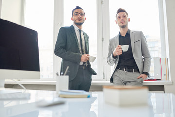 Two young white collar workers wearing suits looking away while enjoying fragrant coffee at spacious open plan office