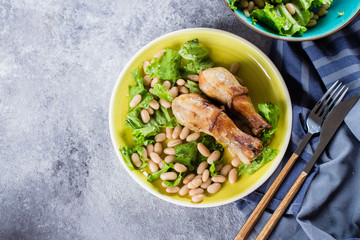 Baked chicken legs with white beans and lettuce leaves on yellow plate on gray stone table background. Food Lunch Dinner Concept.