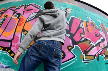 A young guy in a gray hoodie paints graffiti in pink and green colors on a wall in rainy weather. Fisheye shot