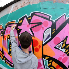 A young guy in a gray hoodie paints graffiti in pink and green colors on a wall in rainy weather. Fisheye shot