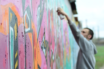 A young guy in a gray hoodie paints graffiti in pink and green colors on a wall in rainy weatherю...