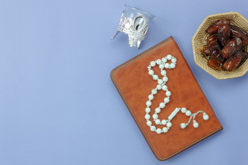 Table top view aerial image of decoration Ramadan Kareem holiday background.Flat lay date in wood basket with white rosary & candle.The holy book of Koran on modern rustic purple paper at office desk.