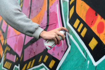 A young guy in a gray hoodie paints graffiti in pink and green colors on a wall in rainy weather