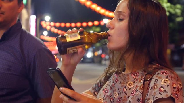 A young girl walks with friends in the night city, drinks beer, flips through the phone. 4k, 3840x2160, HD