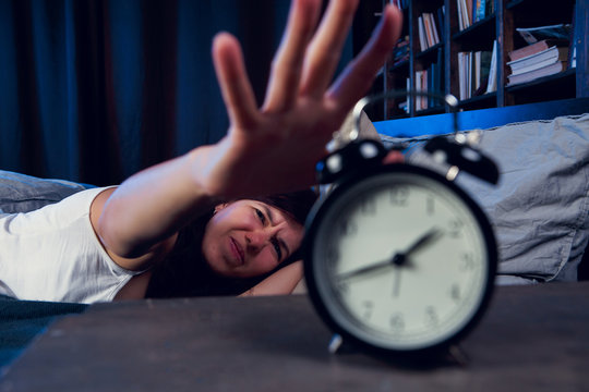 Photo of dissatisfied brunette with insomnia stretching arm to alarm clock at night