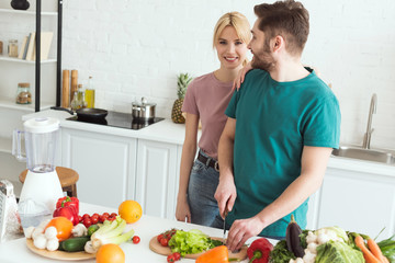 vegan girlfriend hugging boyfriend while he cutting vegetables at kitchen