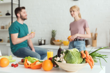 vegan couple talking in kitchen with fruits and vegetables on foreground