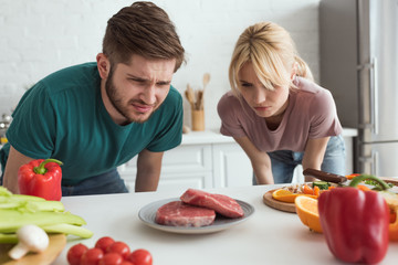disgusted vegan couple looking at raw meat on plate in kitchen at home