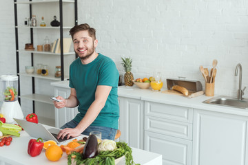 smiling man with credit card sitting at table with laptop in kitchen at home