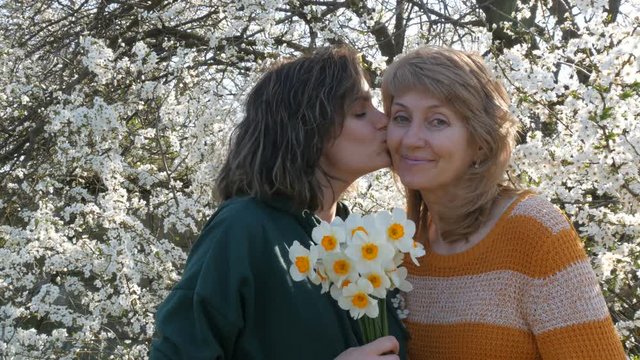 Adult daughter and mother smile each other in the spring against a blossoming tree. The daughter kisses mom in Mothers Day