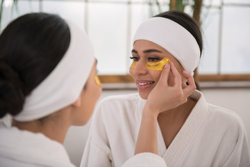 Mutual help. Cheerful young woman looking at her friend while putting an eye patch on her face