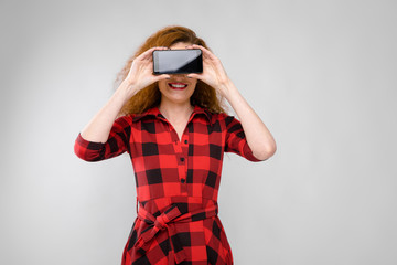 Portrait of beautiful redhead happy young woman in checkered dress smiling holding mobile phone in front of her face on gray background