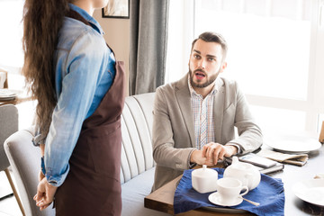Pensive handsome businessman in light gray jacket sitting at table with teapot and cup and asking waitress about lunch while making order in restaurant