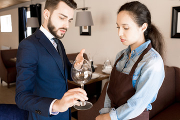 Serious pernickety handsome restaurateur with beard pointing at wineglass and lecturing upset waitress in apron in high quality restauran