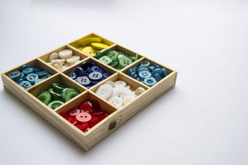 Clothes buttons in wooden box on white cardboard background. Closeup photo.