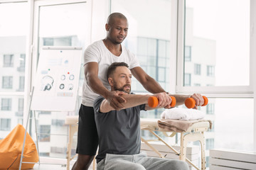 Exercising equipment. Nice pleasant serious man sitting on the medball while holding dumbbells in front of him