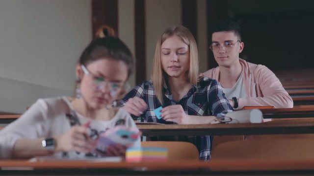 A female student unfolds a note, which was given by a classmate. Cheat sheet, love note. Group of three modern students in lecture theater: guy and two female students.