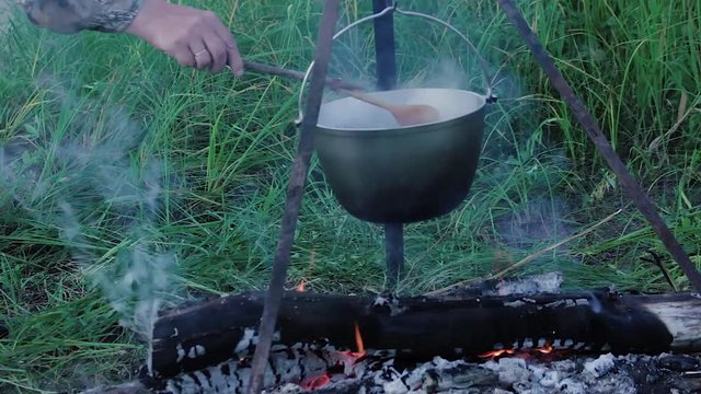 Bowler at the stake The fisherman removes scaling with a wooden spoon in a bowler hat suspended on a tripod over a campfire close up