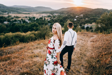 Attractive young loving couple of man in white shirt and with suspenders and gentle girl in dress with red flowers are walking on sunny mountain background in sunset. Hold hands