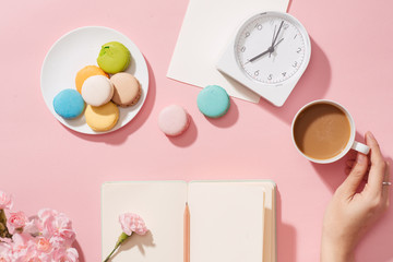 Female hand with coffee cup, macaron, office supply, gift and notebook on pastel desk top view. Fashion pink woman workplace background for blogging. Flat lay. Beautiful morning breakfast.
