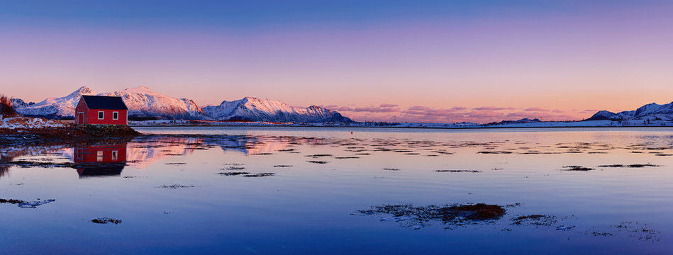 Landscape with beautiful winter lake, red rorbu house and snowy mountains at sunset at Lofoten Islands in Northern Norway. Panoramic view