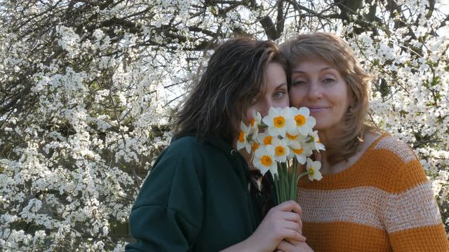 Very beautiful mother age and adult daughter with a bouquet of flowers hugging smiling smiles against the background of a blossoming tree looking at the camera