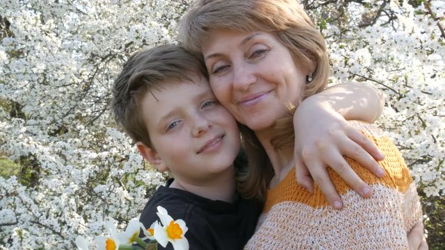 An adult middle-aged mother hugs and kisses her teenager's son tenderly against the background of a beautiful luxuriant tree. Mother's Day in Spring