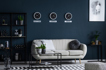 White sofa, coffee table, metal shelf with decorations and three clocks on the grey wall in a living room interior