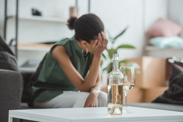 close up shot of glass with wine bottle on table and frustrated young woman sitting in armchair