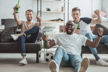 young happy multicultural male friends watching football match and celebrating with beer bottles in...