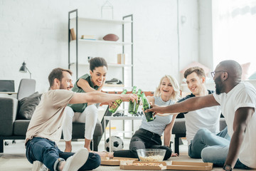 happy multiethnic friends sitting on floor and clinking with bottles of beer