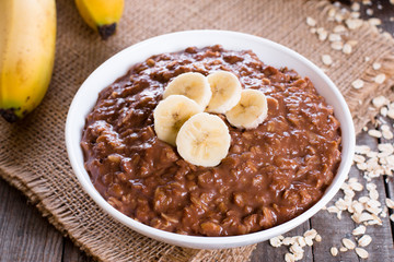 Chocolate oatmeal for breakfast with slices of a ripe banana in a white bowl on a wooden background in a horizontal position.