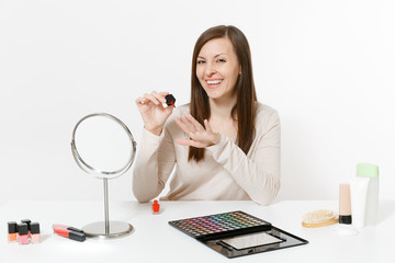 Beautiful young woman painting nails with red nail polish, sitting at table applying makeup with set facial decorative cosmetics isolated on white background. Beauty female fashion lifestyle concept.
