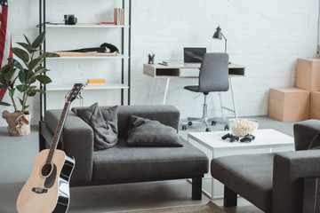 interior of modern living room with armchairs, guitar, shelves and tables with laptop, lamp, popcorn and joysticks