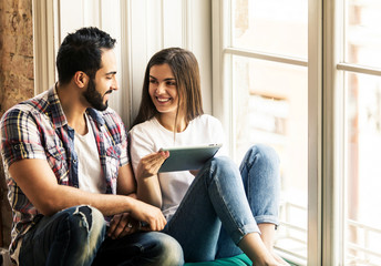 Bearded handsome man and brunette girl sitting on the wide windowsill using tablet computer