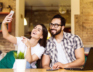Beautiful couple, bearded man and brunette girl taking selfie with smartphone in modern coworking office
