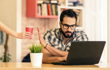 Concentrated freelance worker, bearded handsome man working with laptop and refusing cup of coffee...