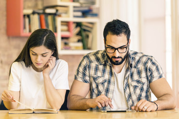 Couple of students get knowledge from book and tablet computer sitting in modern stylish room with bookshelves on background