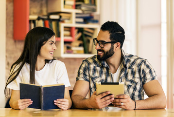 Couple of students hold book and tablet computer sitting in modern stylish room with bookshelves on background