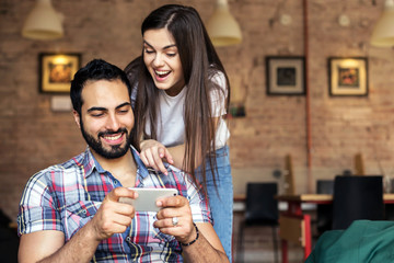 Bearded handsome man and brunette girl having fun playing online game on smartphone in stylish room, happy emotions concept