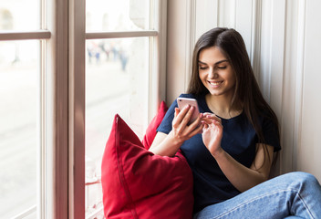 Sexy brunette girl wears blue t-shirt holds smartphone sitting on wide windowsill with big red pillow