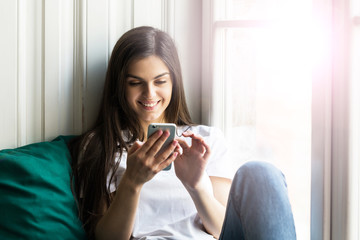 Beautiful smiling brunette girl wears white t-shirt using smartphone sitting on the windwsill